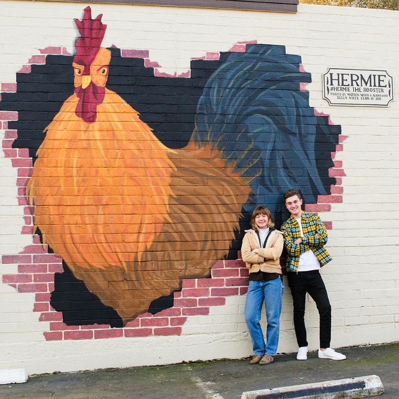 man and woman standing next to a mural of a rooster