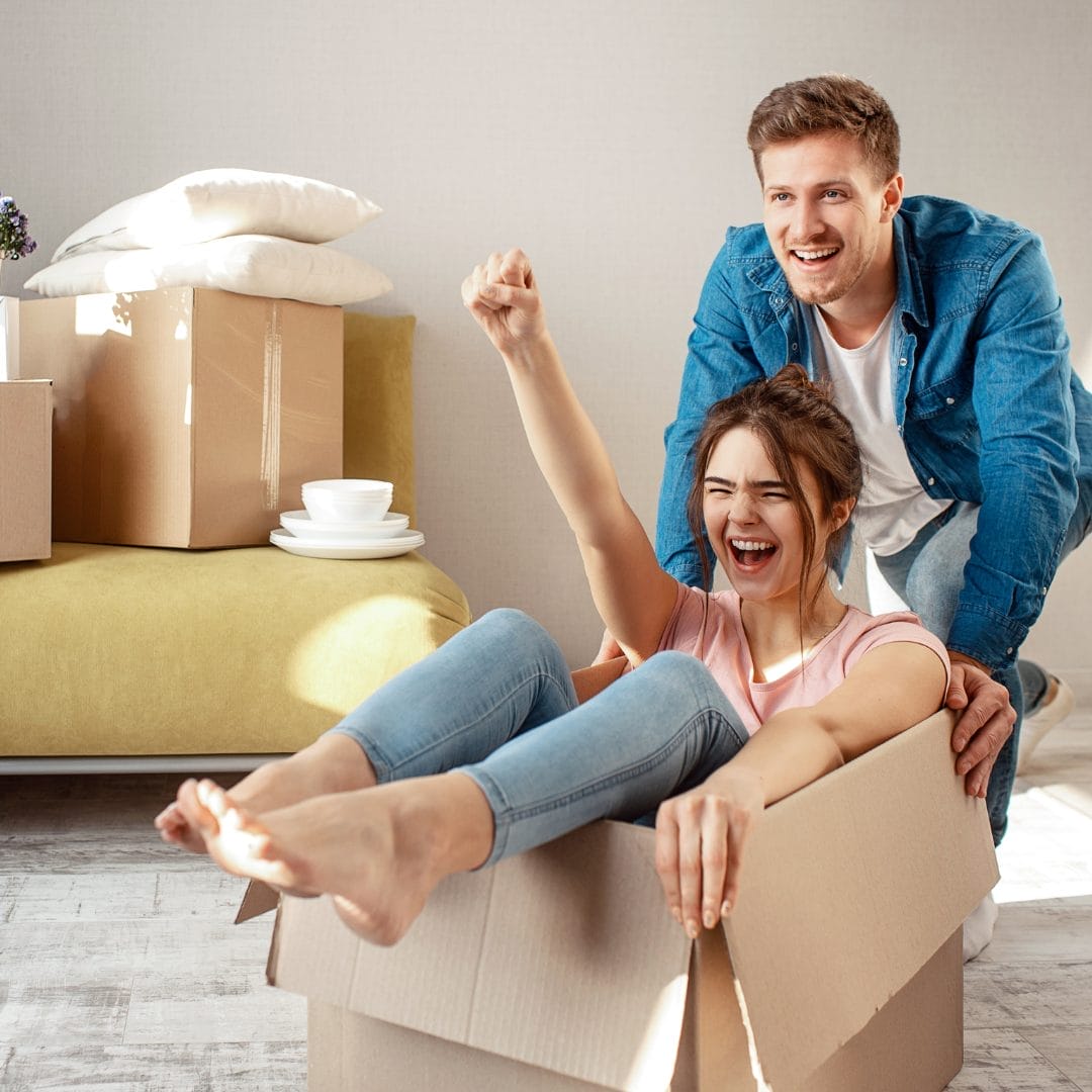 young man and woman excited to move into their first home, photo has him pushing a moving box that she is riding in.